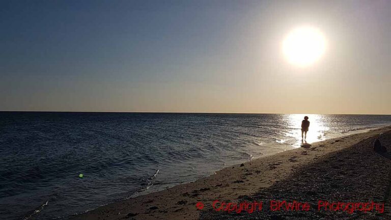 A lone person standing in the sun reflections along the sea on the beach